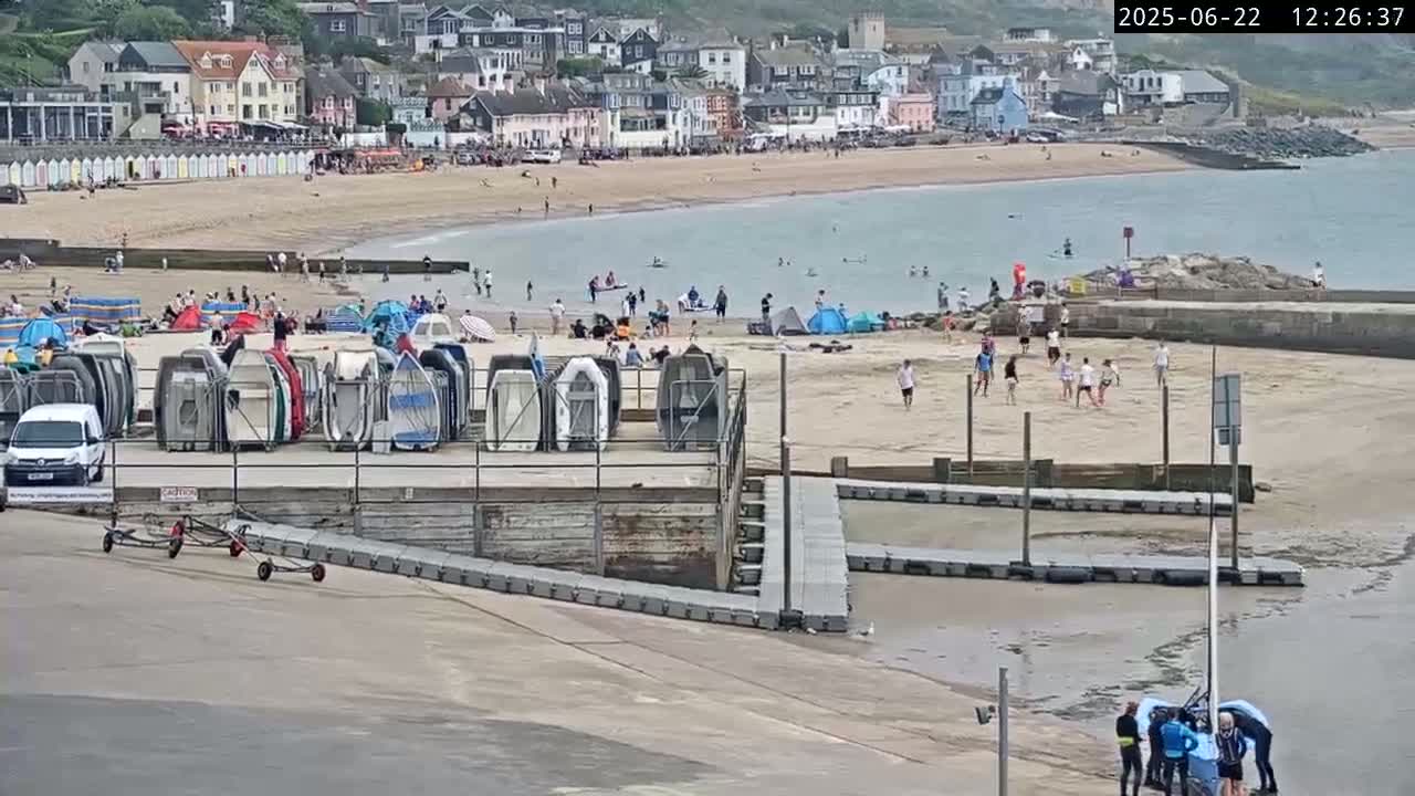A sandy beach with many people, numerous upturned boats on a nearby dock, and a calm body of water is viewed from above on a mostly sunny day.