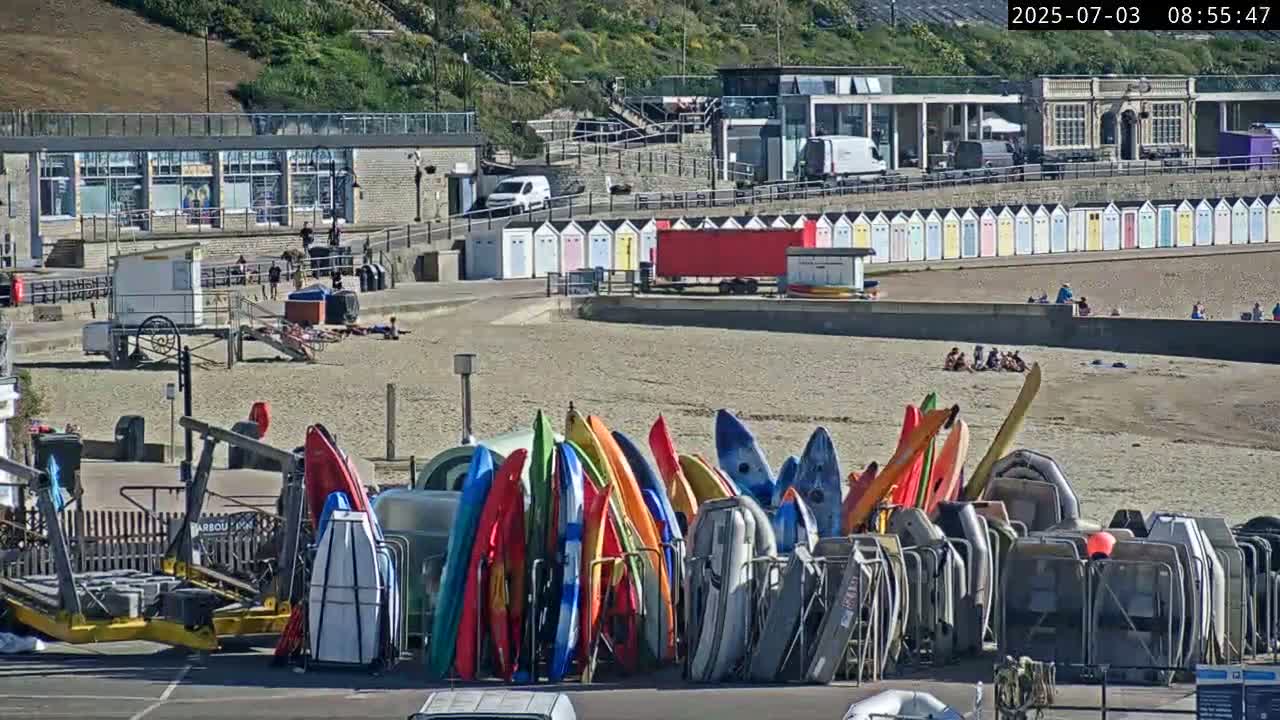 A sandy beach with colorful kayaks and beach huts, under sunny skies, is seen next to a building with a parking lot.