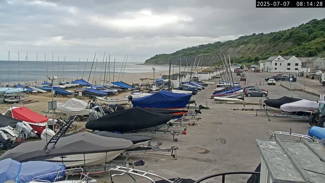 Numerous boats, many covered, are parked in a lot next to a beach and a seaside cliff under an overcast sky.