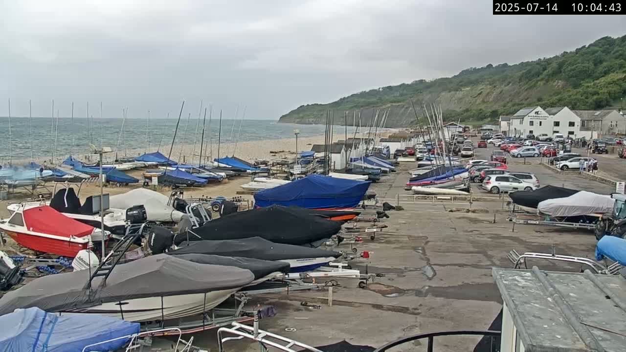 Numerous boats, many covered, are parked in a lot next to a sandy beach and a seaside cliff under an overcast sky.