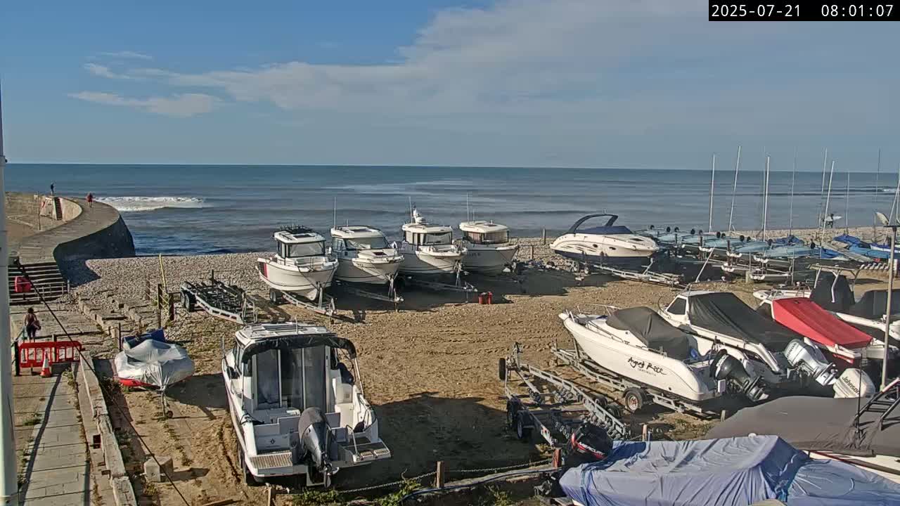 Several motorboats are docked on a pebble beach next to a calm, blue ocean under a partly cloudy sky.