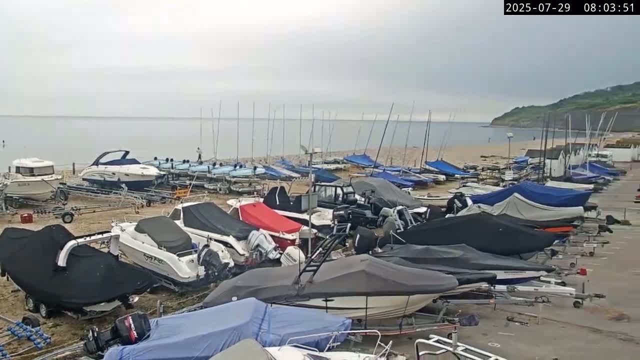 Numerous boats, many covered, are parked along a sandy shore next to calm, grey water under an overcast sky.