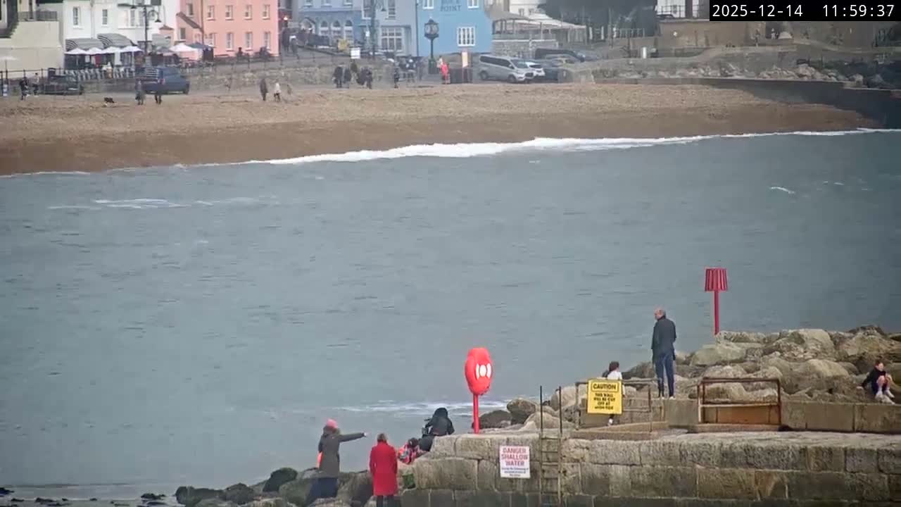 Small boats are moored in a harbor next to a stone pier featuring a cannon, under a grey and overcast sky.