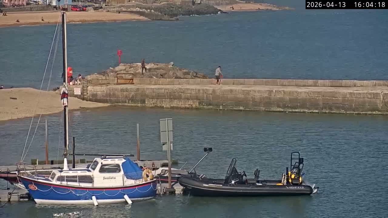 Small boats are moored in a harbor next to a stone pier featuring a cannon, under a grey and overcast sky.