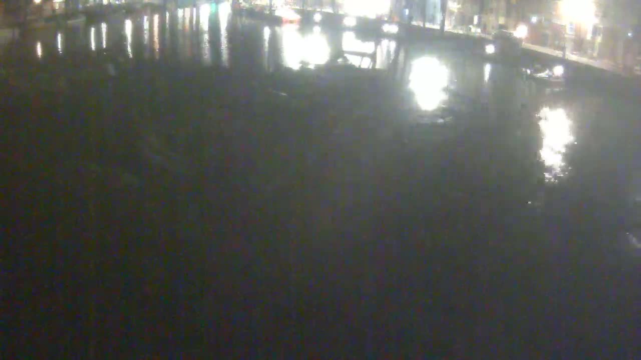 The dark, still water of a canal reflects the lights of buildings and vehicles on the banks at night.