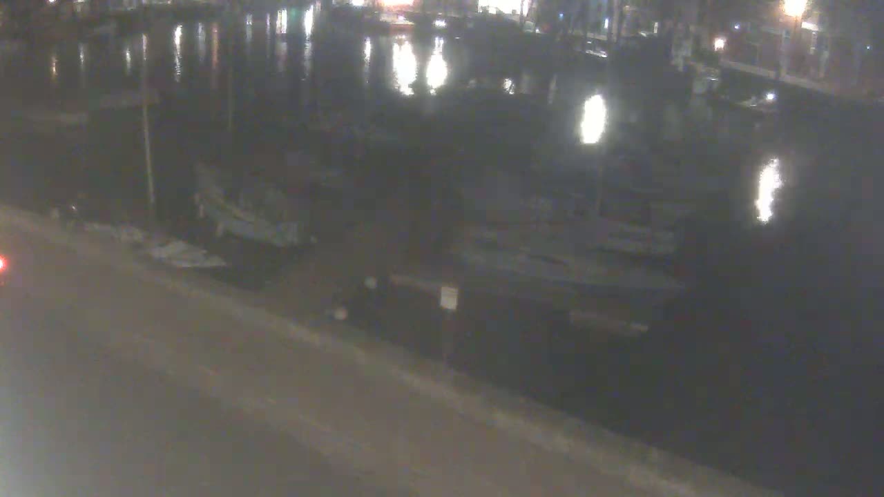 At night, several boats are moored along a calm body of water next to a road.