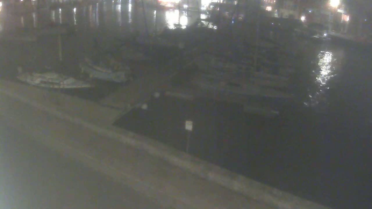 Several boats are moored at a dock at night, beside calm water reflecting the lights from a nearby town.