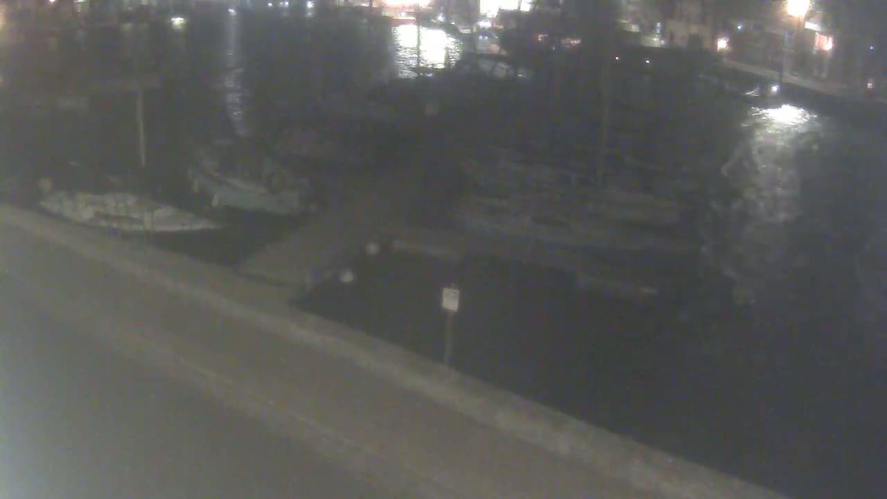 At night, several boats are moored along a dark waterfront, with a calm body of water reflecting the lights from the distant buildings.