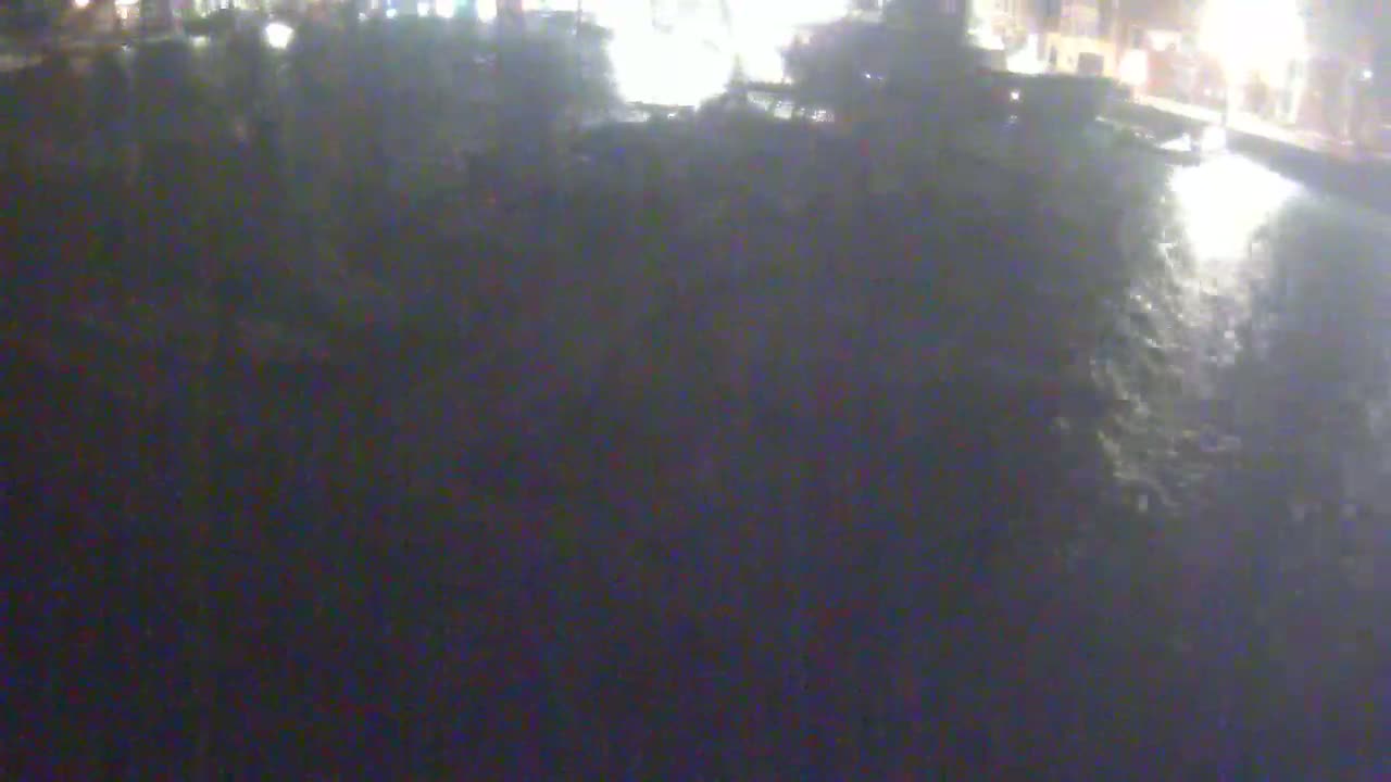 A dark, nighttime scene shows a blurry view of a body of water reflecting lights from a distant structure.