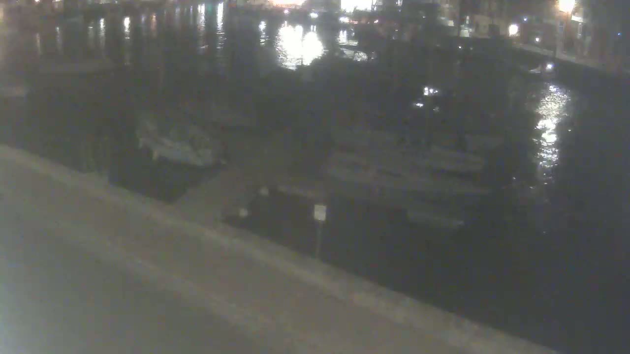 A nighttime view shows several boats moored along a dark waterway, with a paved area visible in the foreground.
