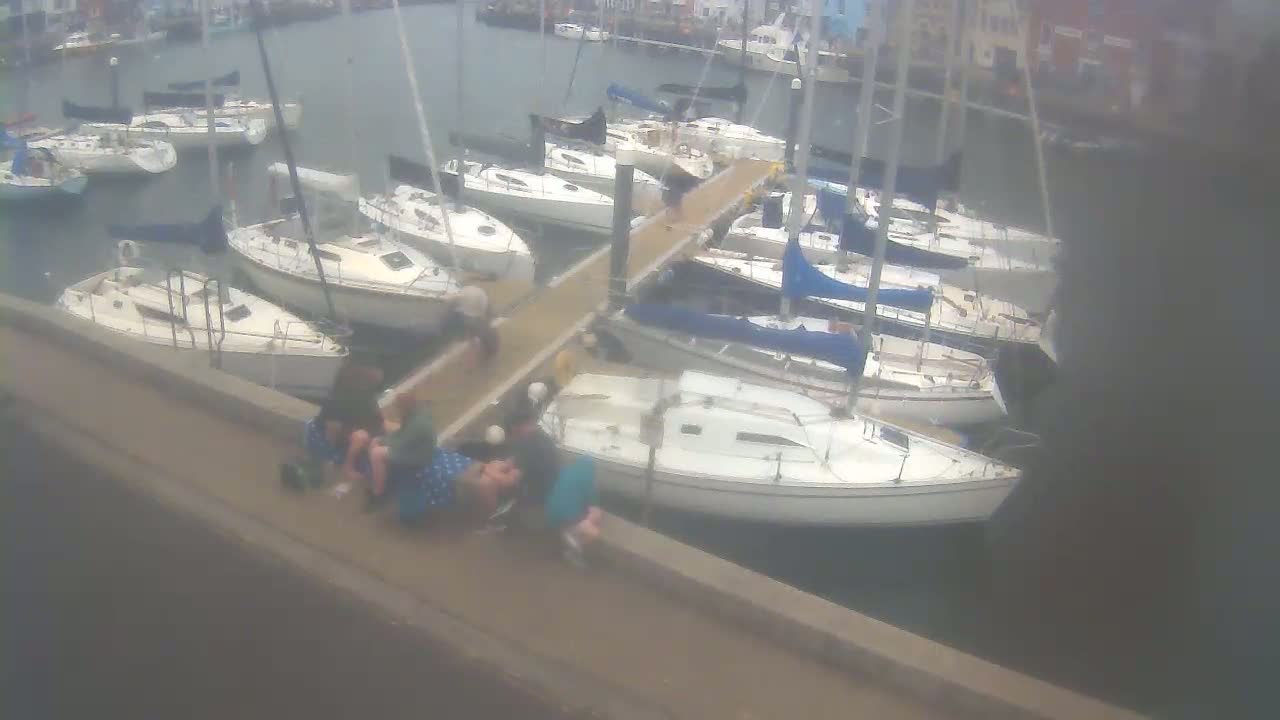 Several sailboats are docked at a marina on an overcast day, with a small group of people sitting on a nearby wall.