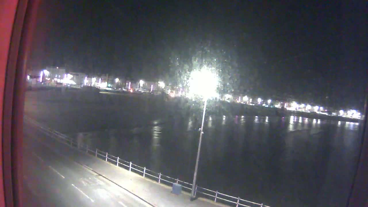 A nighttime view from a window shows a dark body of water reflecting lights from buildings across a road and a promenade.