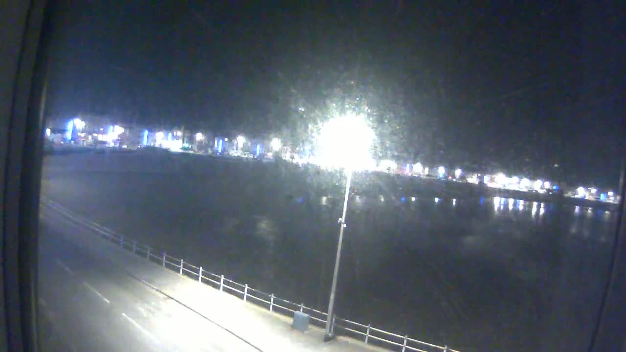 A nighttime view of a calm body of water reflecting city lights, with a road and railing visible in the foreground.