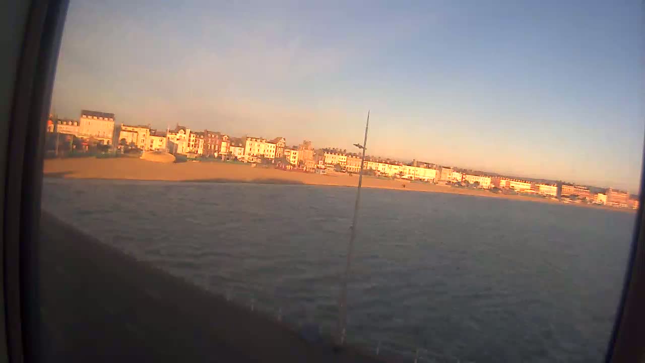 A row of buildings sits along a sandy beach next to a body of water with small waves under a clear, sunset sky.