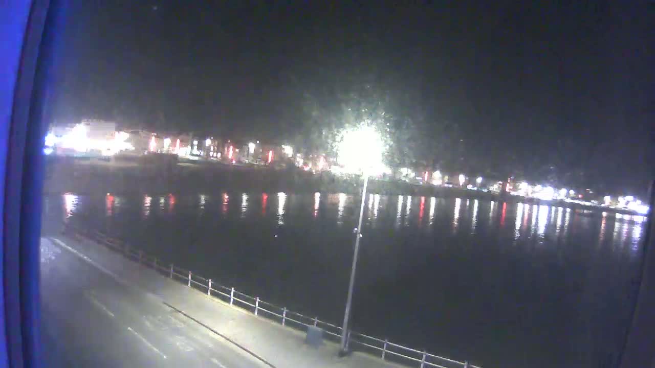 A nighttime view of a calm body of water reflecting the lights of a city across the waterfront, with a paved walkway and railing visible in the foreground.