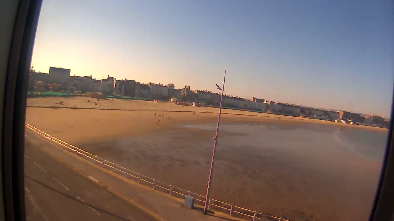 A mostly clear, sunny day reveals a wide sandy beach with a few people scattered across it and buildings lining the shore in the distance.