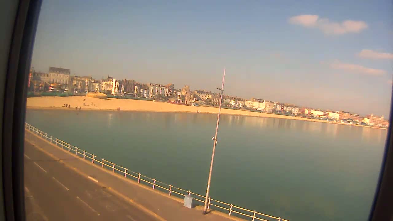 A sandy beach with a few people on it is lined with buildings, next to calm, greenish-blue water under a mostly clear, sunny sky.