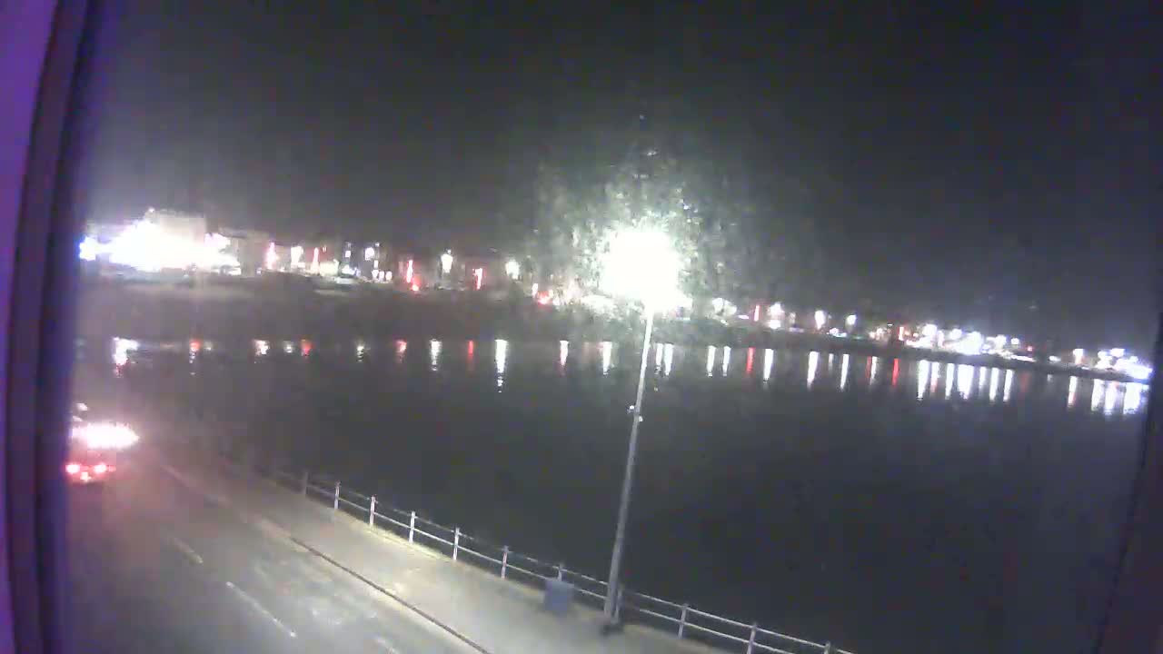 A nighttime view of a calm body of water reflecting city lights, with a road and a vehicle visible in the foreground.
