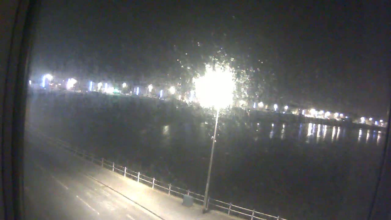 A nighttime view from a window shows a calm body of water reflecting lights from a distant shoreline, with a street and a fence visible in the foreground.