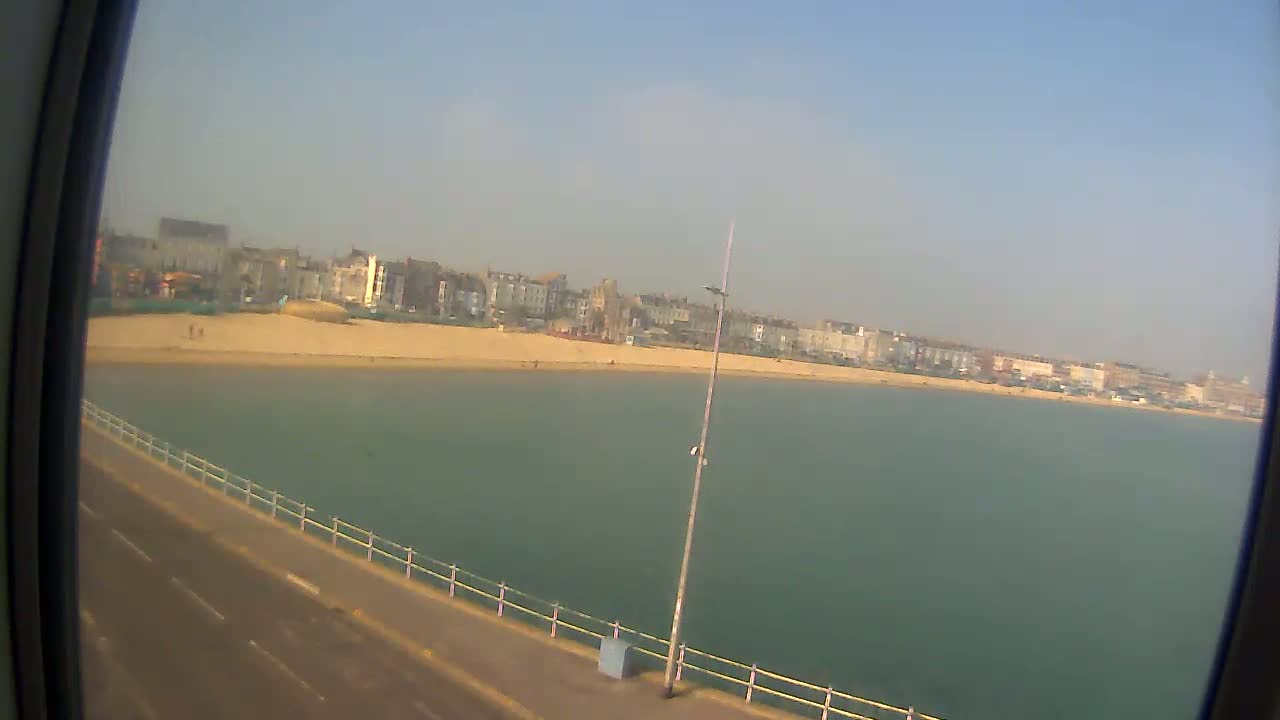 A sandy beach with buildings in the background is visible from a waterfront road on a clear, sunny day.