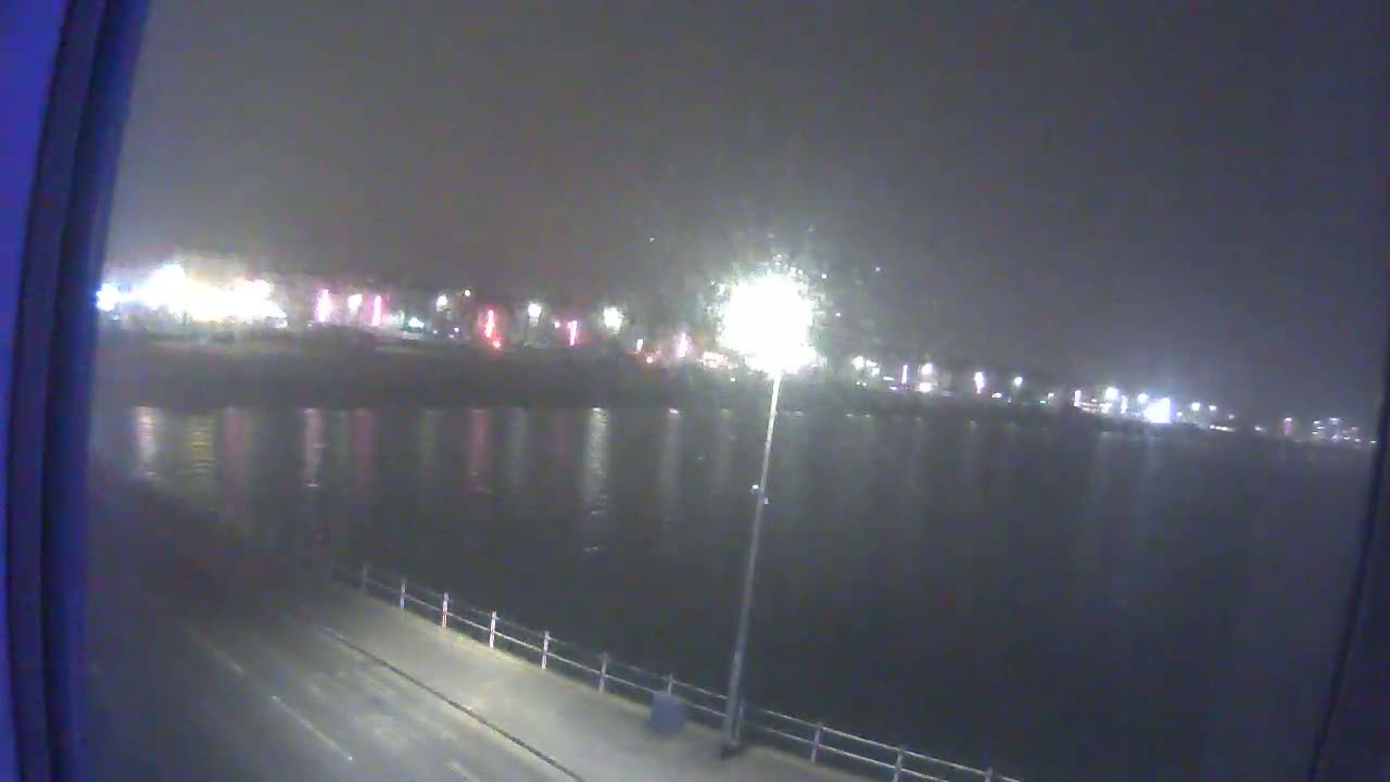 A nighttime view of a calm body of water reflecting the lights of a distant town under hazy conditions.