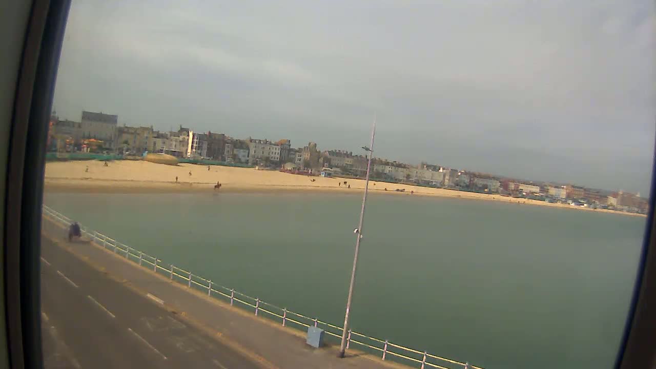 A sandy beach with a few people on it stretches along a calm, green-tinged body of water, backed by a row of buildings under a mostly cloudy sky.