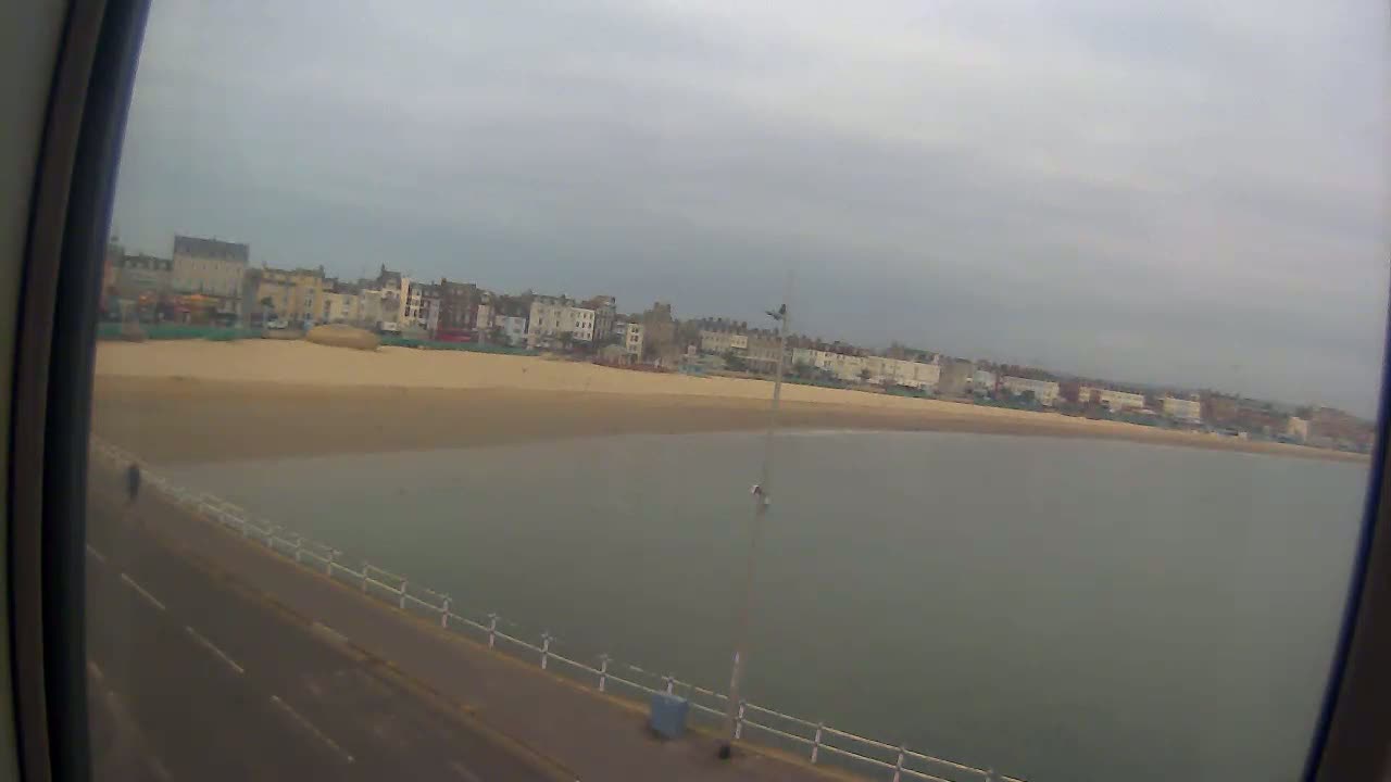 A sandy beach with calm, gray water stretches out before a row of buildings under an overcast sky.