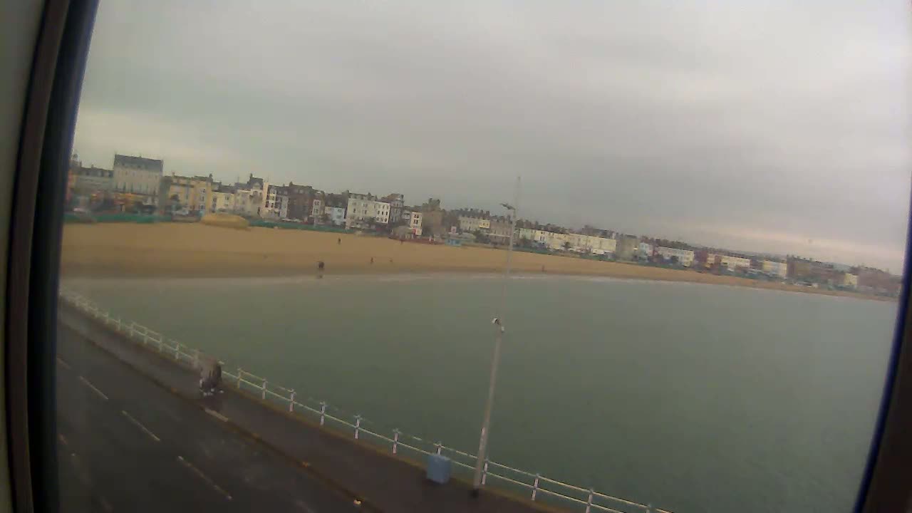 A sandy beach stretches along a row of buildings under an overcast sky, with calm water lapping at the shore.