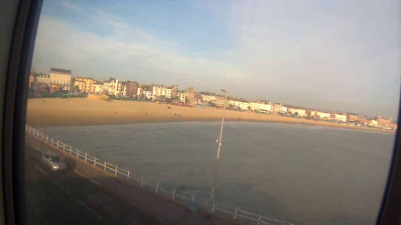 A sandy beach with a calm sea and a row of buildings in the background under a mostly clear sky.