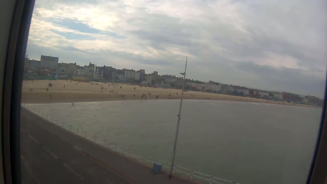 A mostly cloudy day shows a sandy beach with people on it, a calm ocean, and buildings in the background.