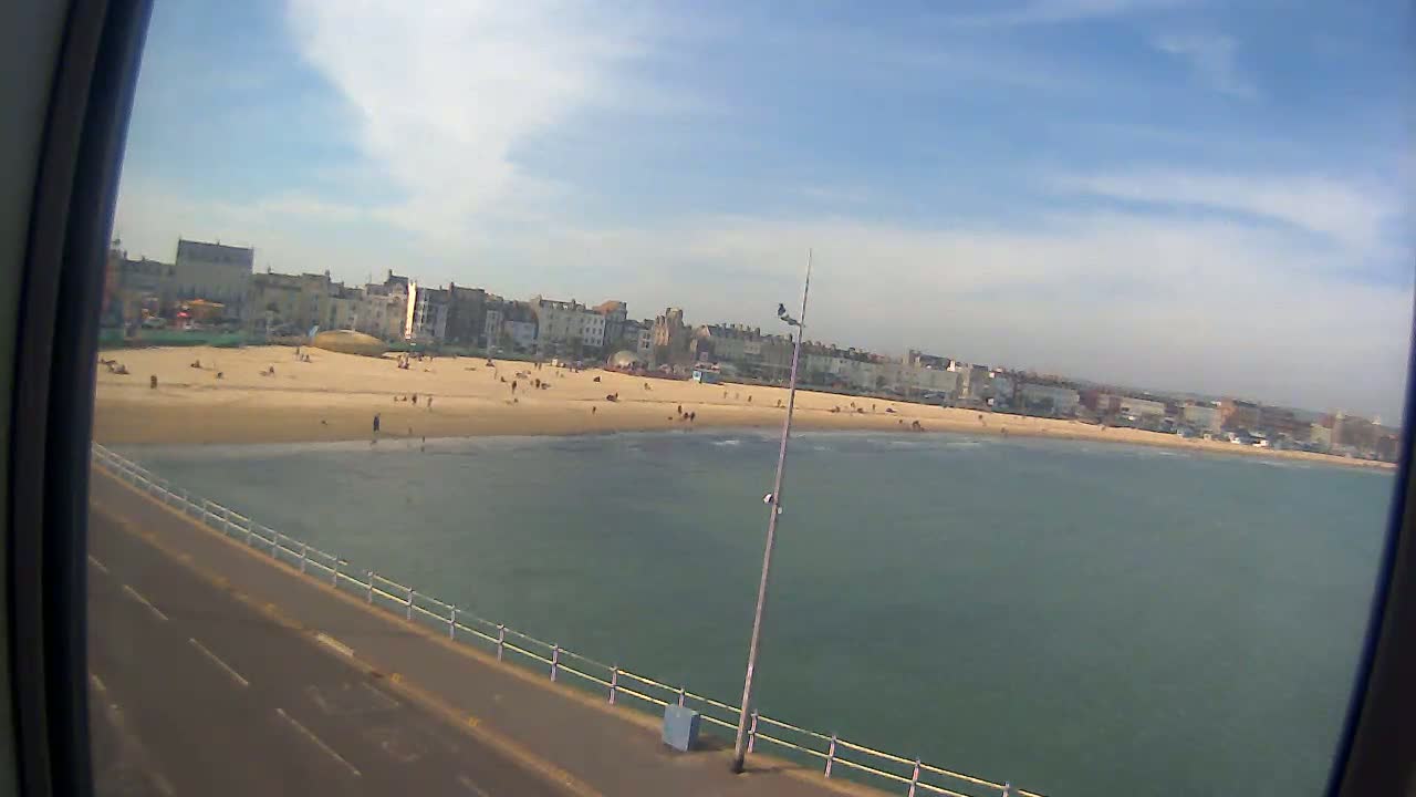 A sandy beach with a moderate number of people is visible, adjacent to a calm, greenish-blue sea under a mostly sunny sky.