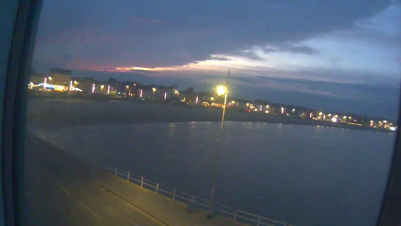A nighttime view of a calm body of water with a lit-up beachfront and a partly cloudy sky.