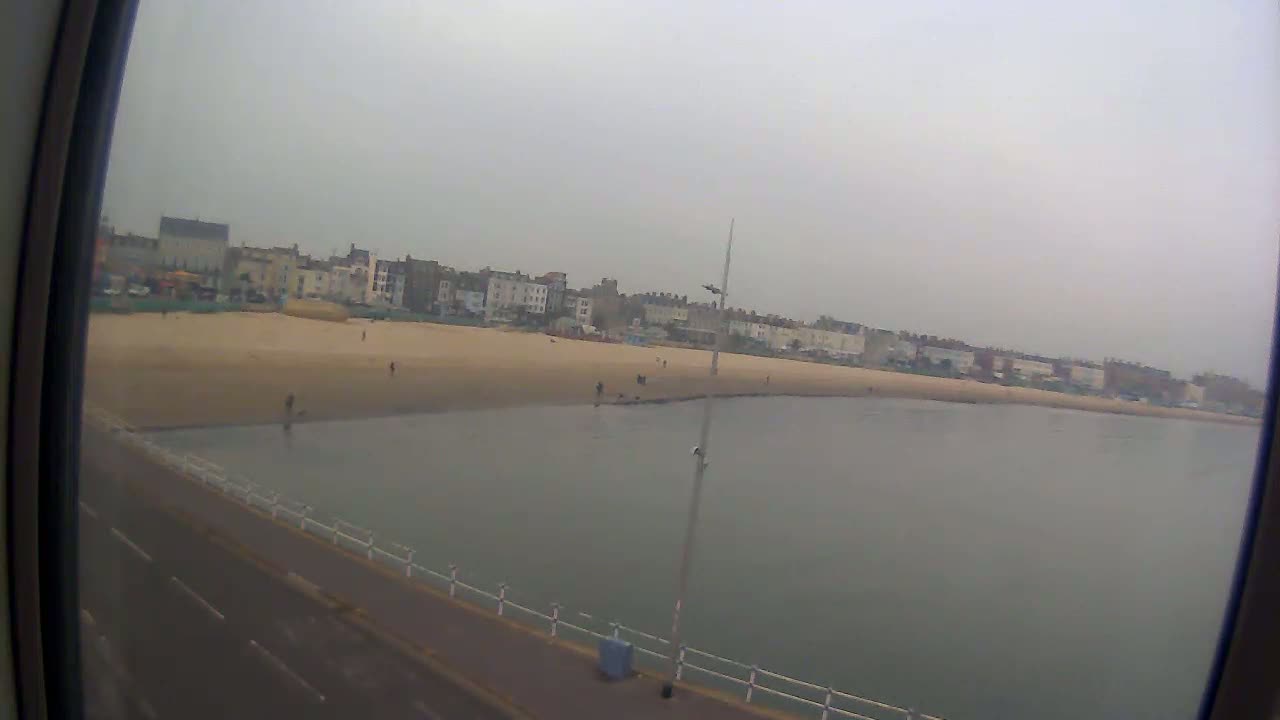 A sandy beach with a few people on it is next to calm, gray water, and a row of buildings stretches across the background under an overcast sky.