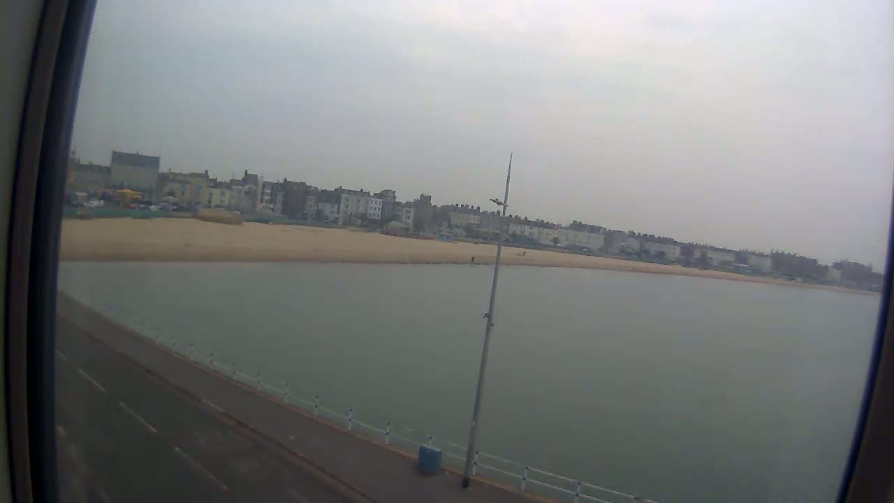 A view from a window shows a calm body of water next to a sandy beach and a row of buildings on an overcast day.