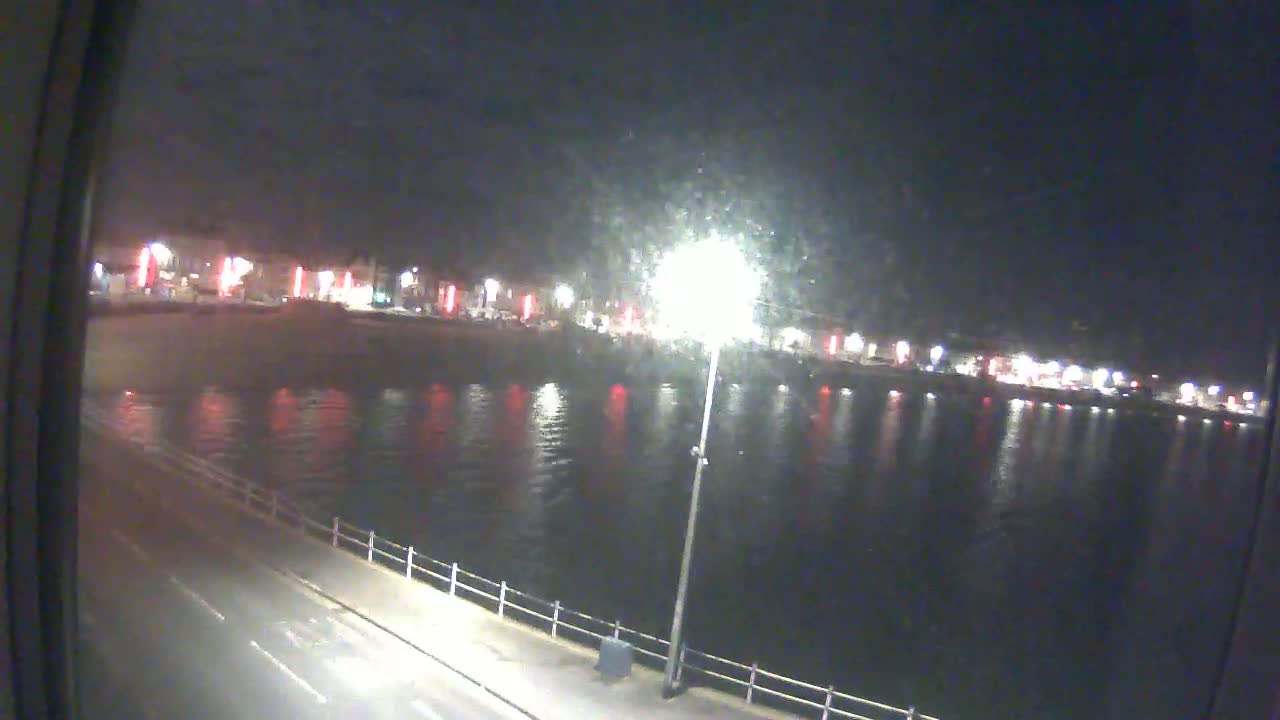 A nighttime view of a calm body of water reflecting lights from buildings across a road.