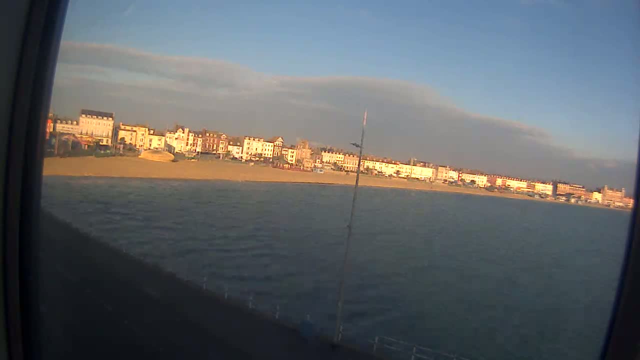A row of buildings lines a sandy beach next to a calm body of water under a mostly clear sky.