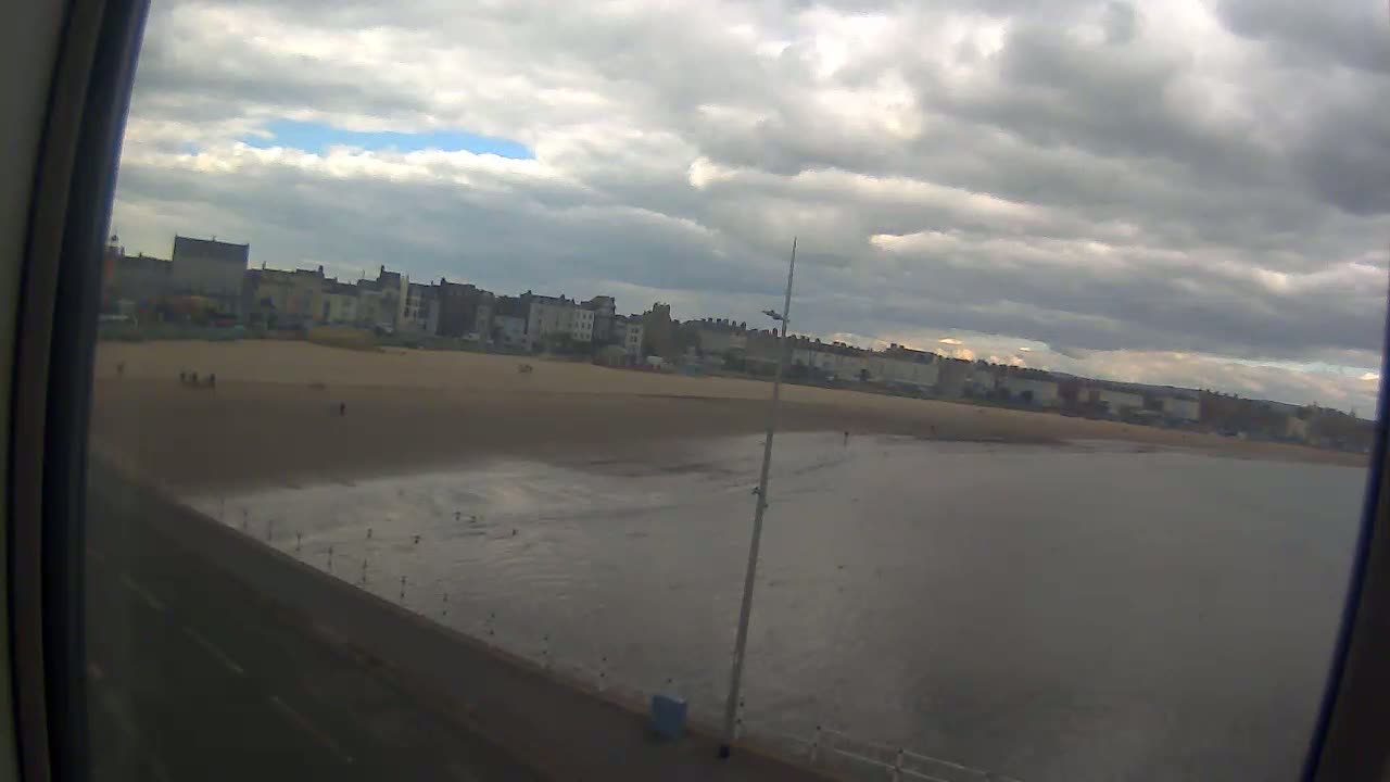 A wide shot from a window shows a sandy beach with a calm sea under an overcast sky, backed by a row of buildings.