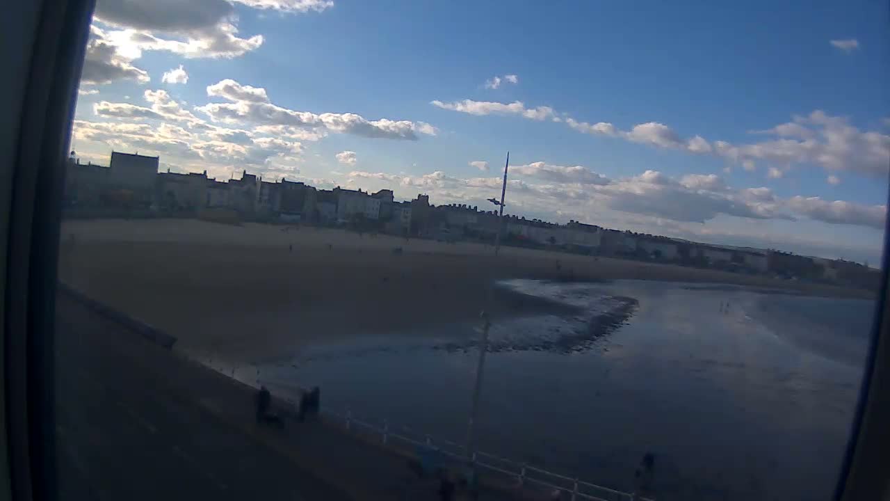 A mostly sunny day reveals a sandy beach with buildings lining the shore and a calm body of water extending to the horizon.