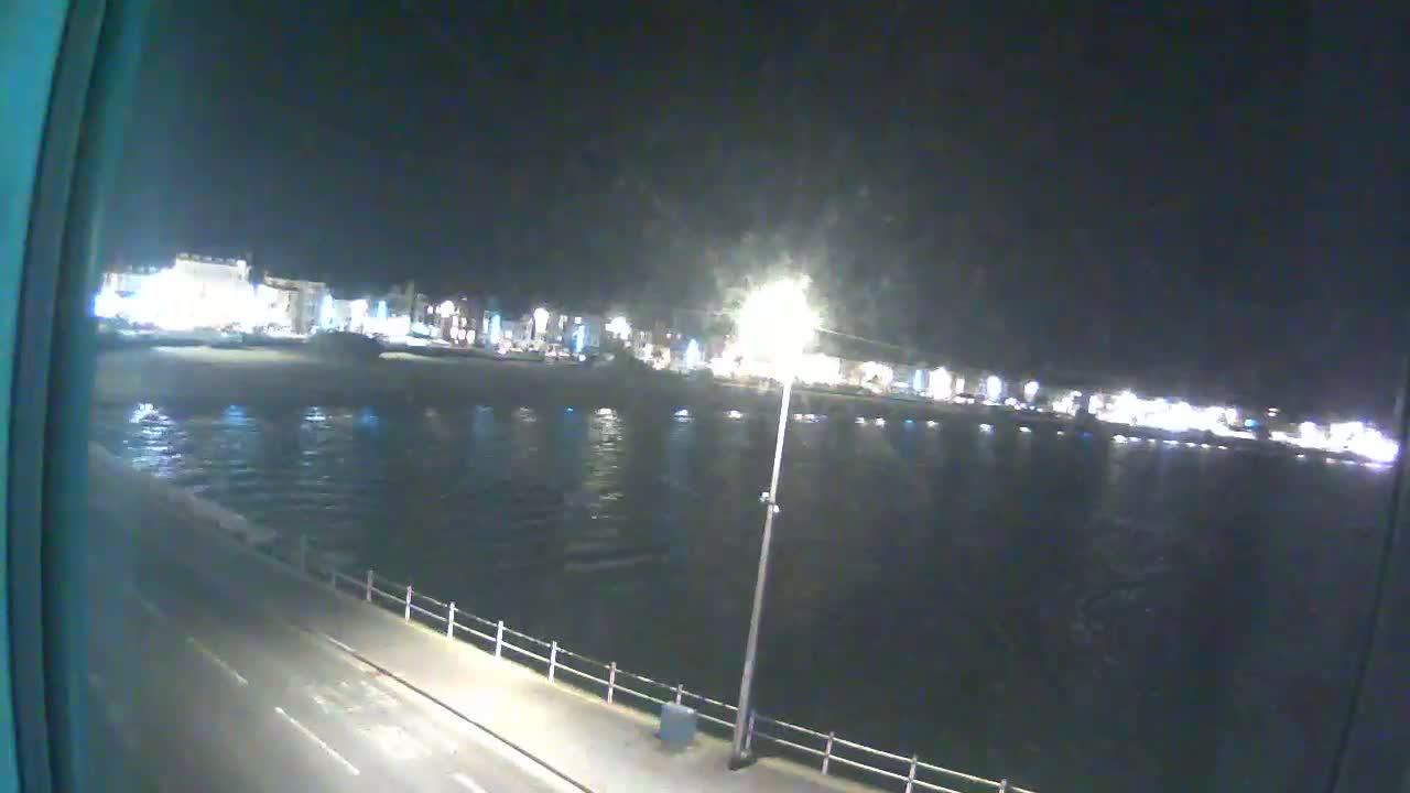 A nighttime view shows a calm body of water reflecting the lights of a town across the shore, viewed from a slightly elevated position near a roadway.