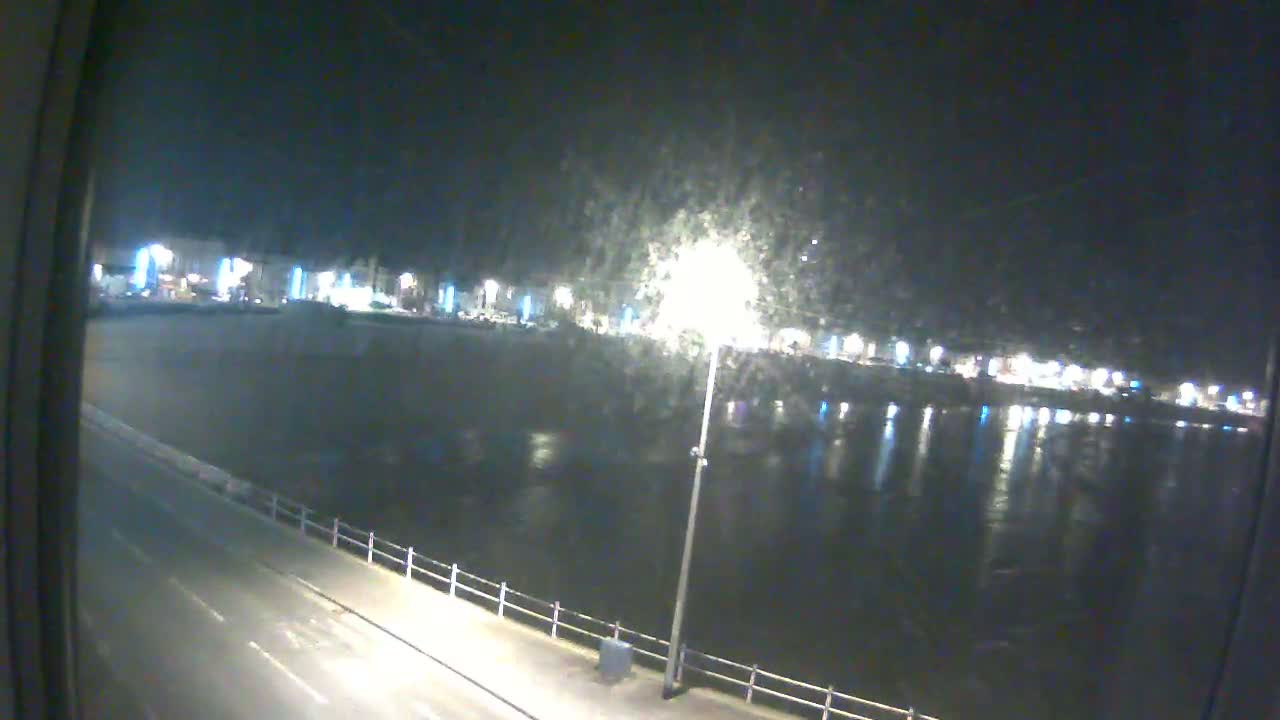 A nighttime view of a calm body of water reflecting the lights of a town across a paved road with a railing.