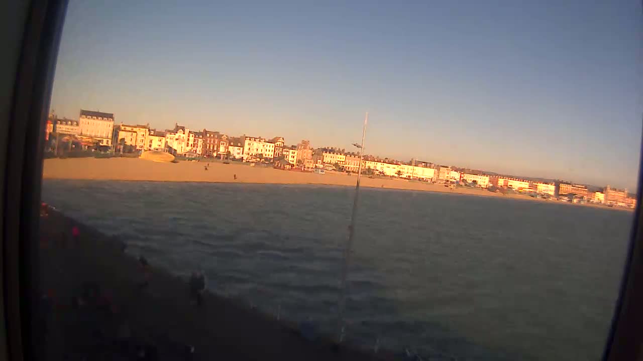 A sandy beach with buildings behind it is visible across a body of slightly choppy water under a clear, light blue sky.