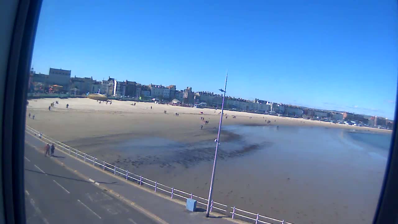 A sandy beach with a few people on it stretches out to calm, shallow water under a clear blue sky.