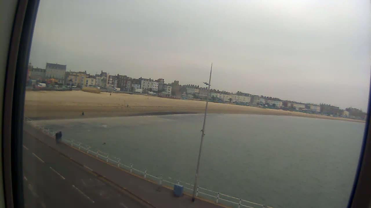 A wide shot from a high vantage point shows a beach with buildings behind it, a calm body of water in front, and a roadway along the water's edge under an overcast sky.