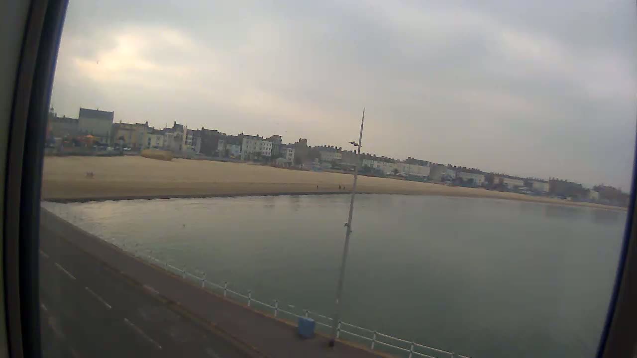 A sandy beach stretches along a calm, gray body of water, backed by a row of buildings under an overcast sky.