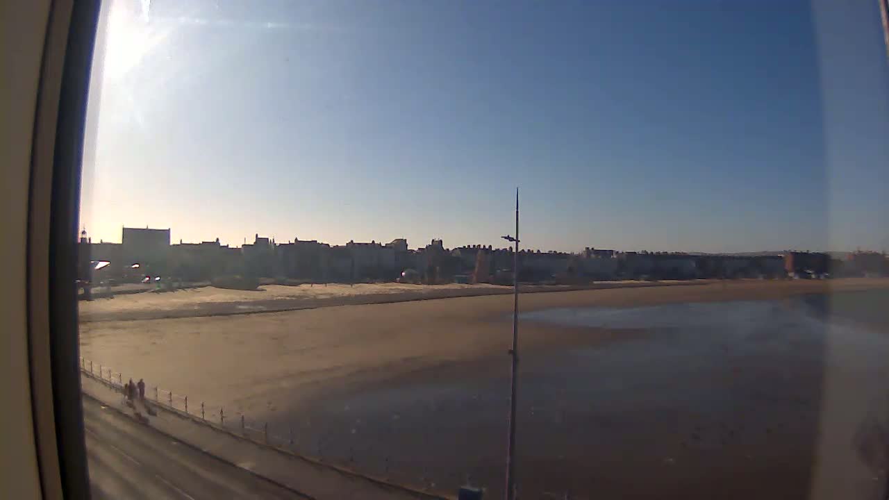 A sandy beach stretches out to a calm body of water, with a row of buildings visible in the background under a clear, sunny sky.