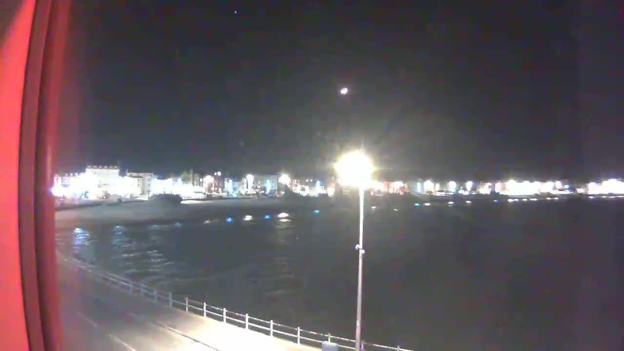 A nighttime view of a calm body of water with a shoreline of buildings and a paved walkway.