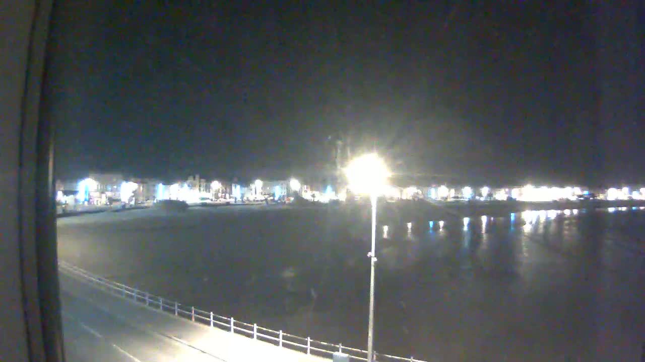 A nighttime view of a calm body of water reflecting the lights of a town across a paved waterfront promenade.