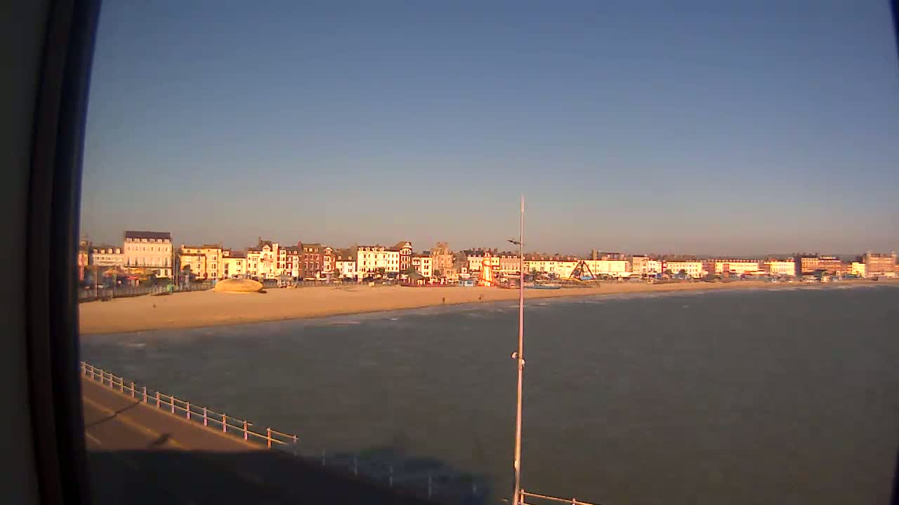 A sandy beach with calm water and a row of buildings behind it is visible under a clear, bright sky.
