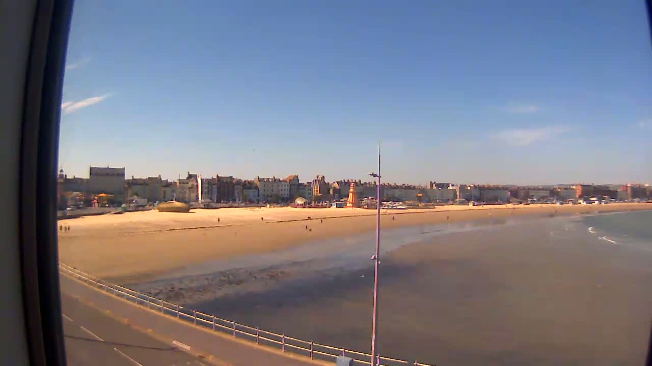 A sandy beach stretches out to a calm sea under a clear blue sky, with a town's buildings visible in the background.