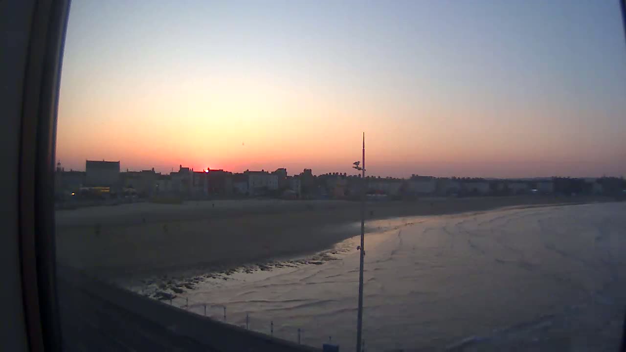 A coastal town at sunset is viewed from a distance, showing a sandy beach and calm ocean waves.
